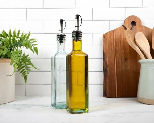 Two glass oil bottles on a kitchen counter with a plant and utensils.